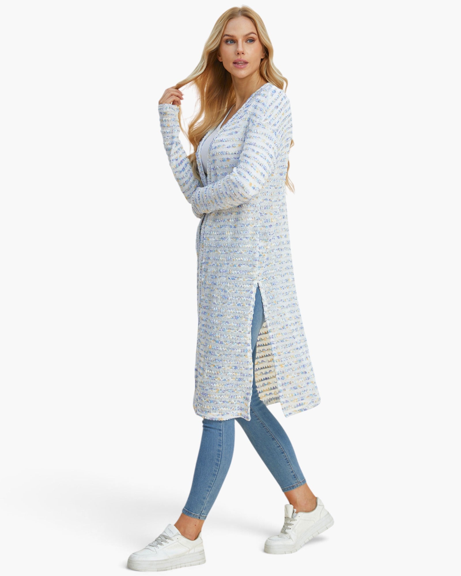A woman modeling a light blue and white textured knit cardigan with a side slit, paired with blue skinny jeans and white sneakers, against a plain white studio background.