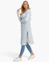 A woman modeling a light blue and white textured knit cardigan with a side slit, paired with blue skinny jeans and white sneakers, against a plain white studio background.