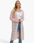 A blonde woman modeling a long, light pink open-front cardigan with a textured knit pattern over a white tank top and blue jeans, set against a plain white studio background.