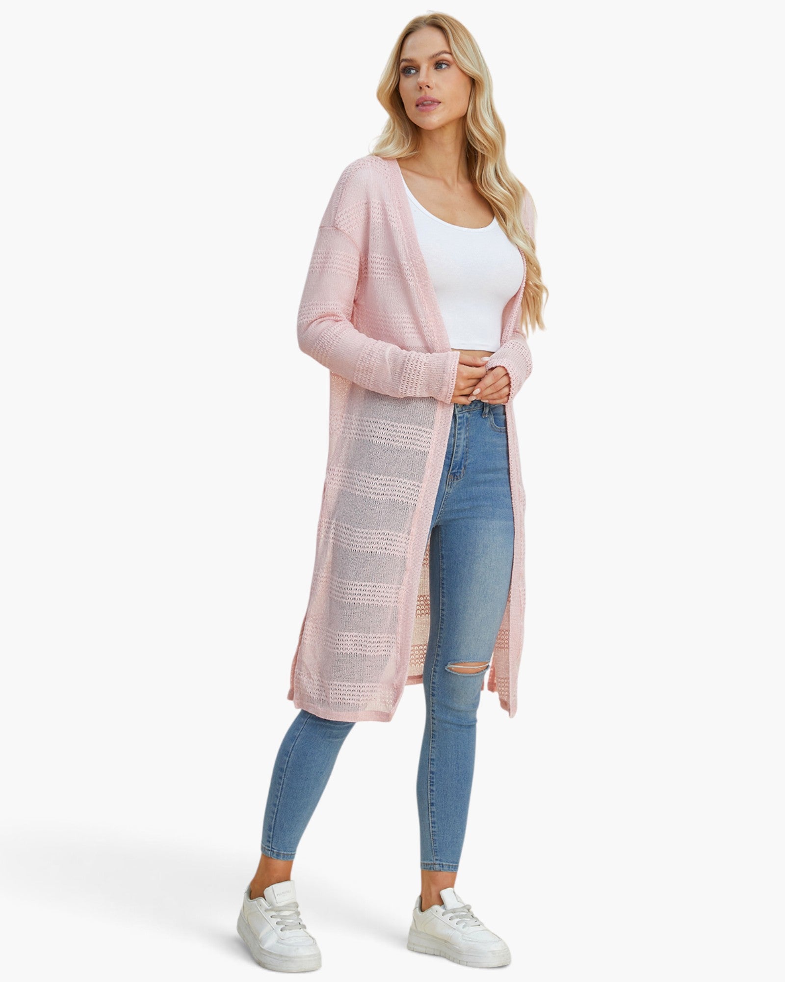 A woman modeling a long, light pink open-front cardigan over a white tank top and blue skinny jeans, standing against a clean white studio background.