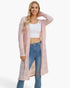 A woman modeling a long, light pink open-front knit cardigan over a white crop top and blue jeans, photographed against a clean white studio background.