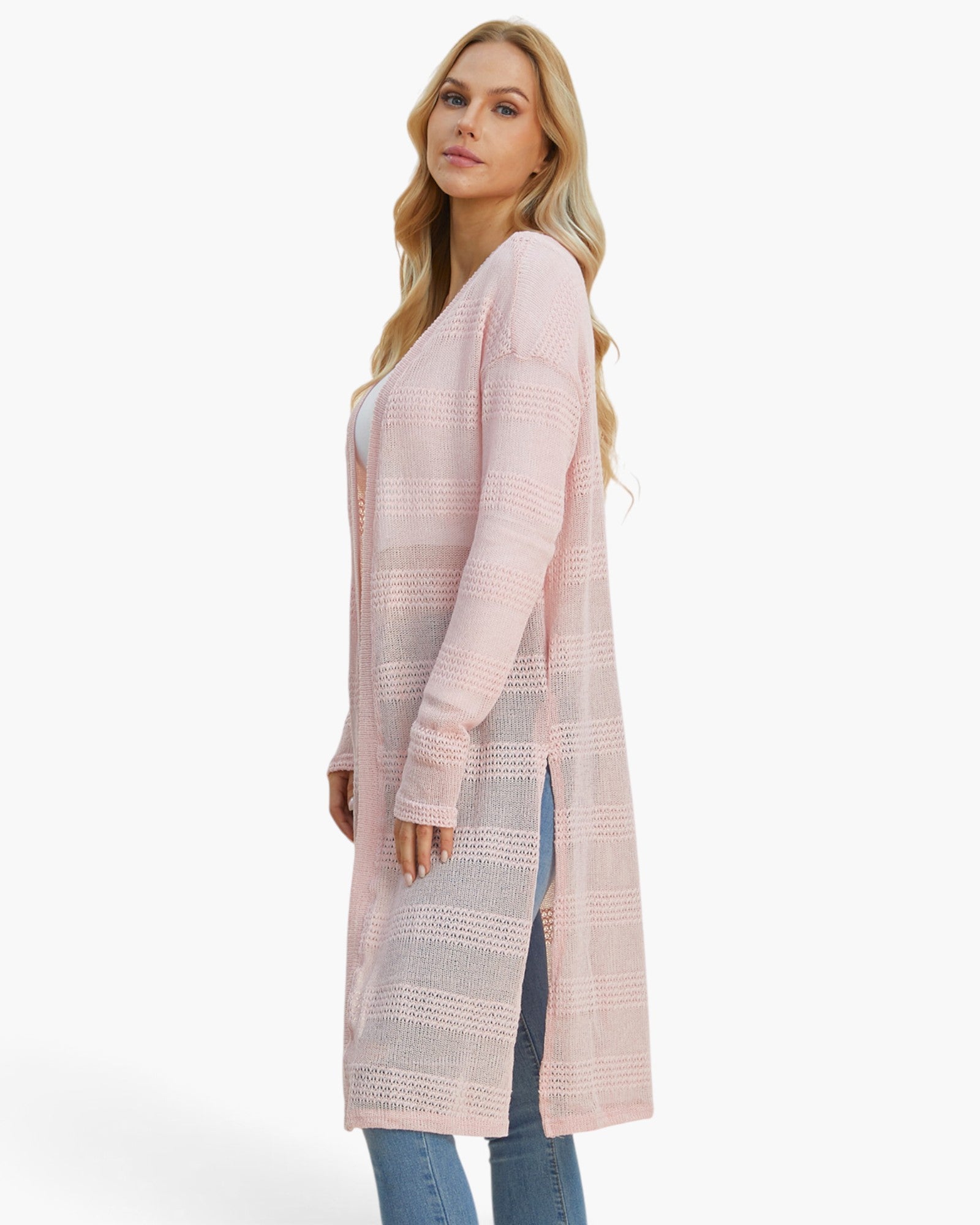 A woman modeling a long, light pink open-front cardigan with a textured knit pattern and side slits, paired with blue jeans against a plain white studio background.