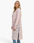 A woman modeling a long, light pink open-front cardigan with a textured knit pattern and side slits, paired with blue jeans against a plain white studio background.