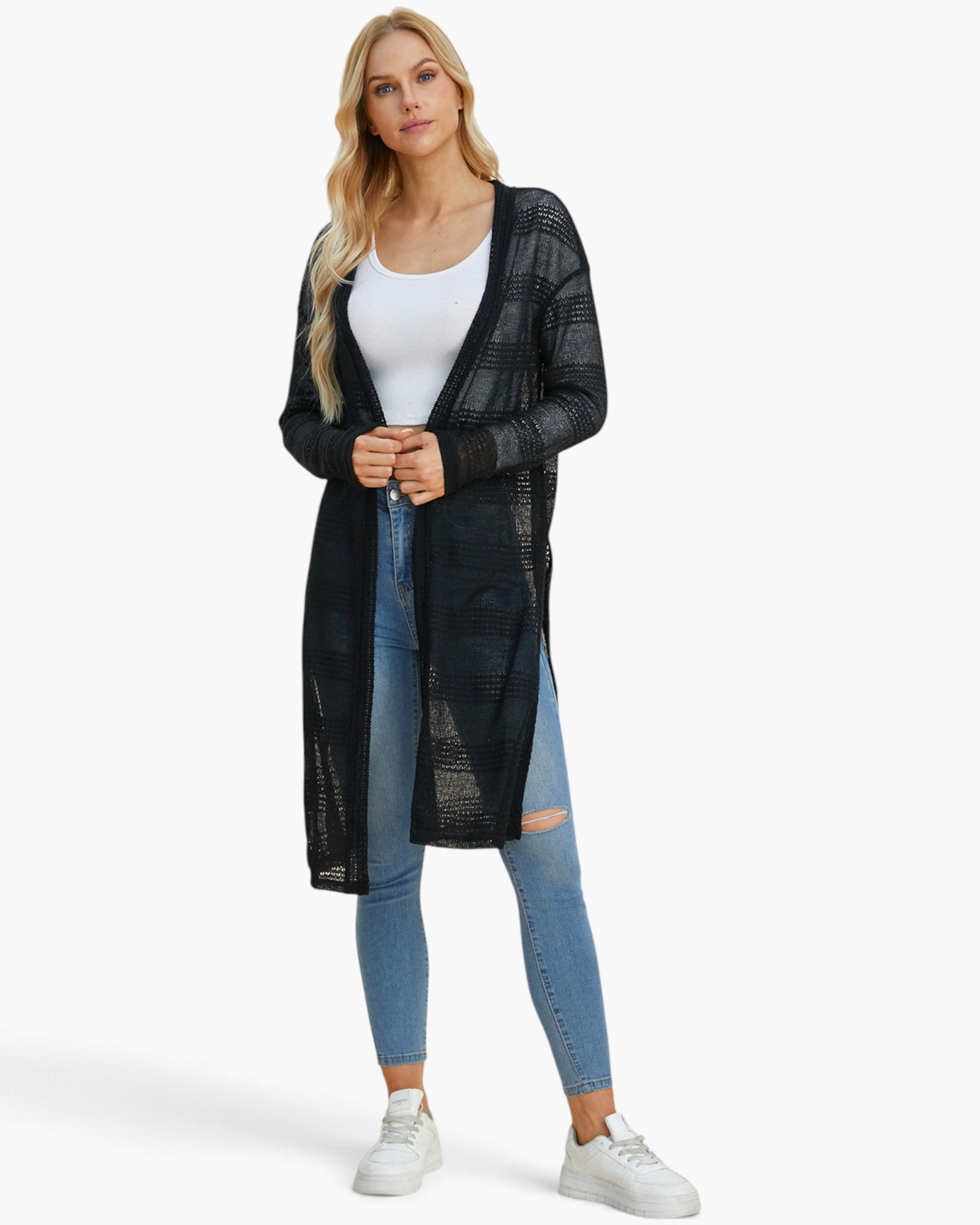 A woman modeling a long, open-front black knit cardigan with a subtle plaid pattern over a white tank top, blue jeans, and white sneakers, set against a plain white studio background.