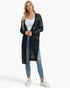 A woman modeling a long, open-front black knit cardigan with a subtle plaid pattern over a white tank top, blue jeans, and white sneakers, set against a plain white studio background.