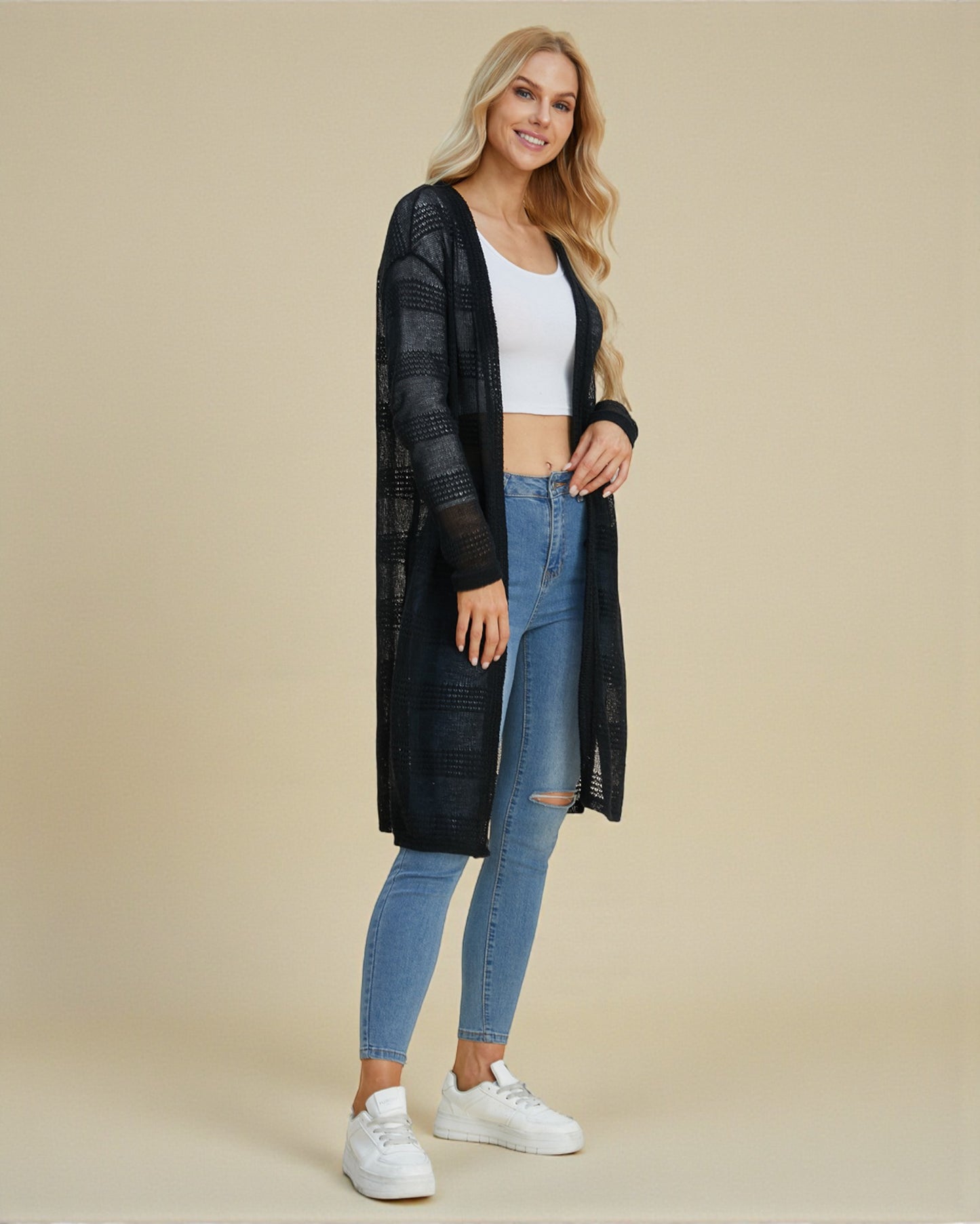Woman wearing a long black cardigan, white top, blue jeans, and white sneakers on a beige background