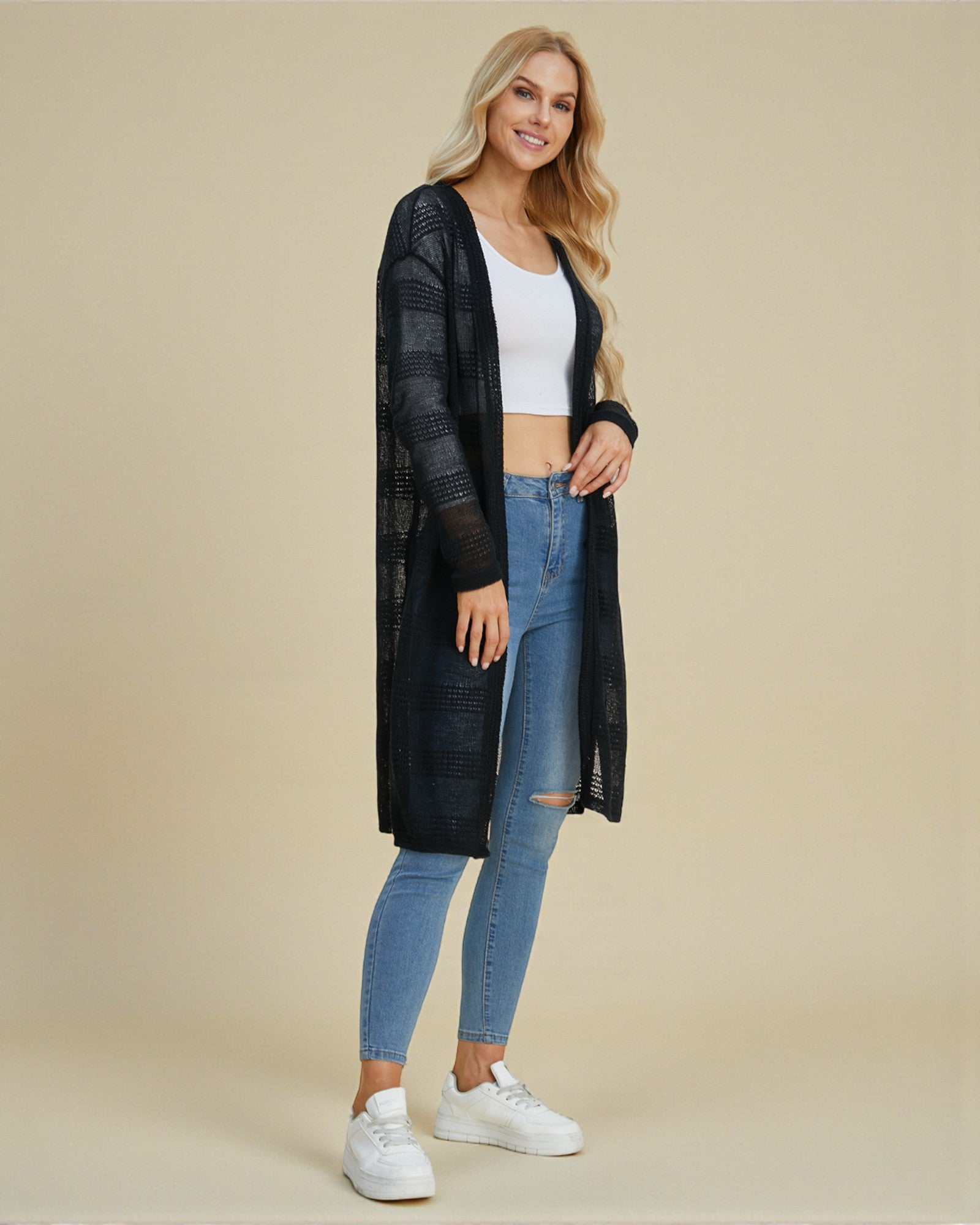 Woman wearing a long black cardigan, white top, blue jeans, and white sneakers on a beige background