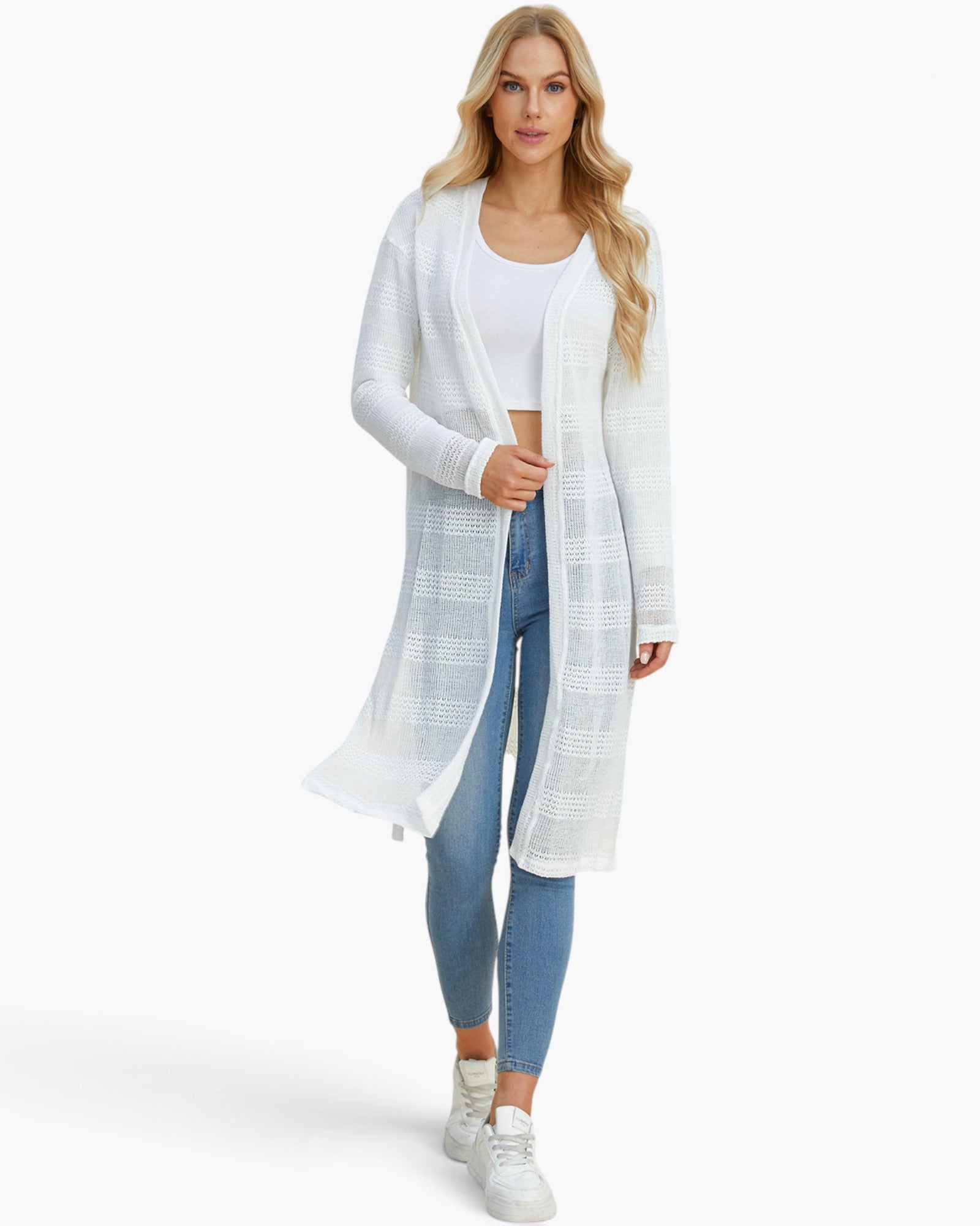 A woman modeling a long white open-front cardigan with a subtle textured grid pattern, worn over a white crop top and blue skinny jeans, against a plain white studio background.
