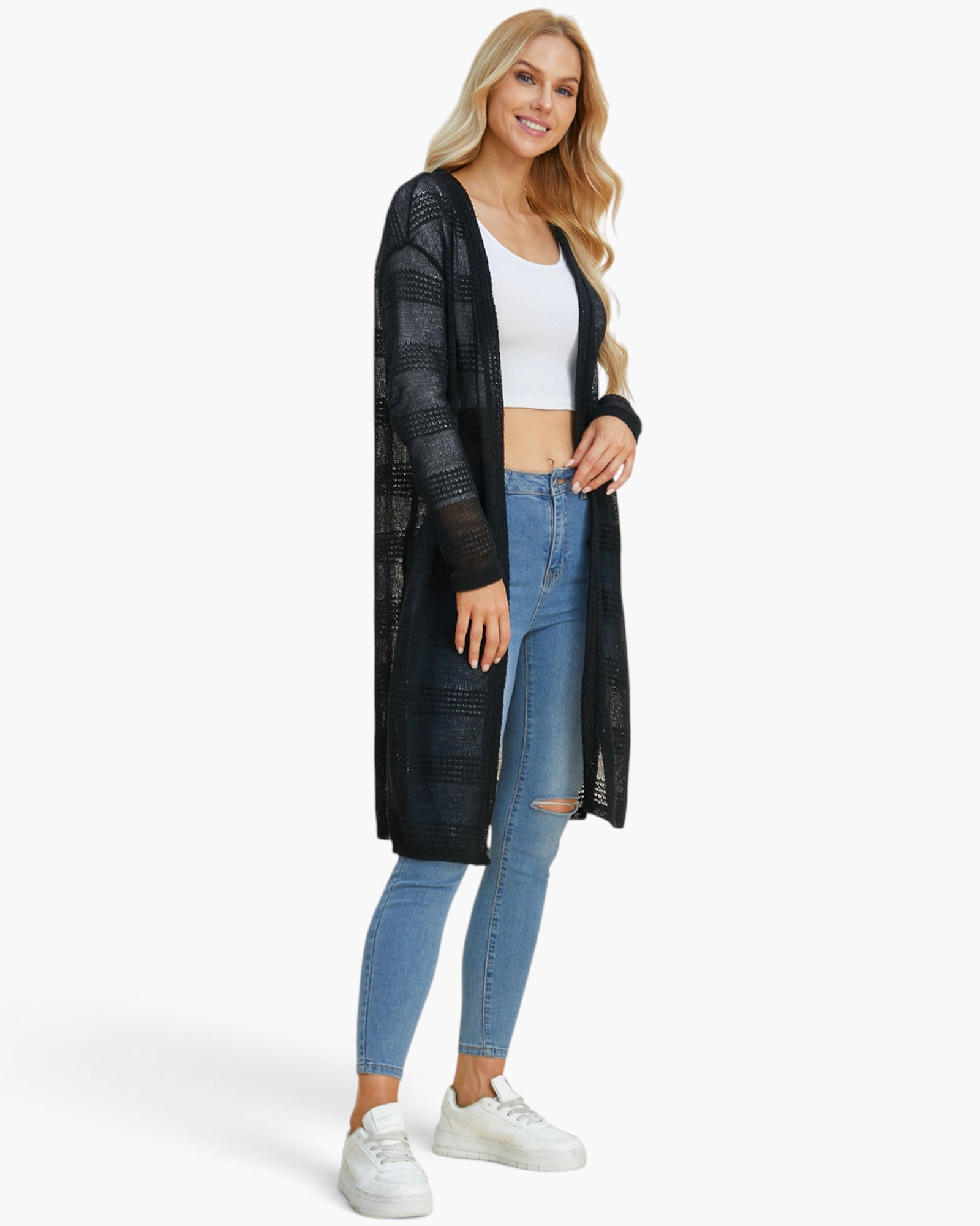A woman modeling a long black open-front knit cardigan over a white crop top and light blue skinny jeans, standing against a plain white studio background.