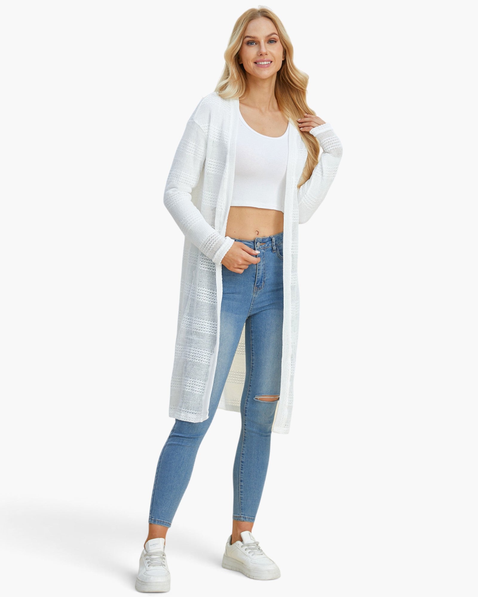 A woman modeling a long white open-front knit cardigan over a white crop top and blue skinny jeans, standing against a plain light gray studio background.