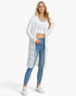 A woman modeling a long white open-front knit cardigan over a white crop top and blue skinny jeans, standing against a plain light gray studio background.