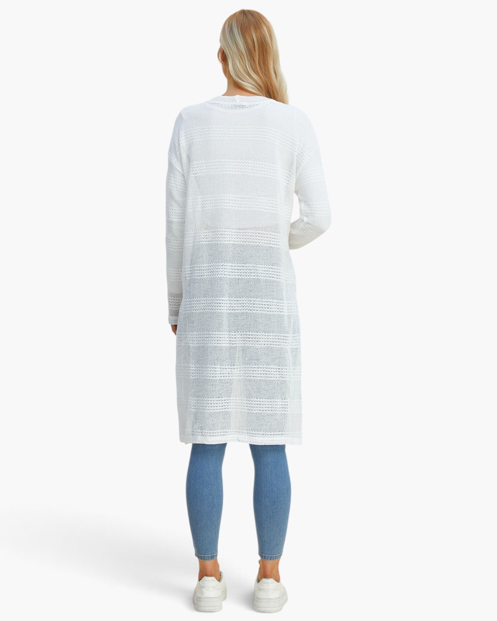 A woman viewed from behind wearing a white long-sleeve knit tunic with horizontal textured stripes, paired with blue leggings and white sneakers, against a plain white studio background.