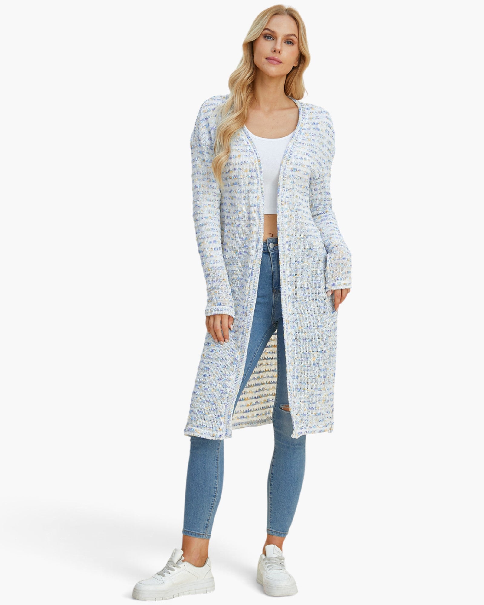 A blonde model wearing a long, open-front knit cardigan with a textured multicolor pattern of light blue, white, and yellow, styled with a white crop top, blue skinny jeans, and white sneakers against a light gray studio background.