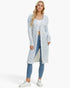 A blonde model wearing a long, open-front knit cardigan with a textured multicolor pattern of light blue, white, and yellow, styled with a white crop top, blue skinny jeans, and white sneakers against a light gray studio background.