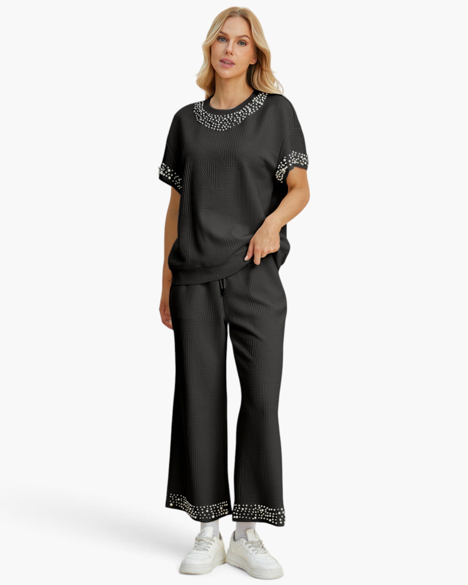 A woman modeling a black two-piece lounge set with white embroidered trim on the neckline and sleeve cuffs, paired with white sneakers against a light gray studio background.