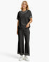 A woman modeling a black two-piece lounge set with white embroidered trim on the neckline and sleeve cuffs, paired with white sneakers against a light gray studio background.