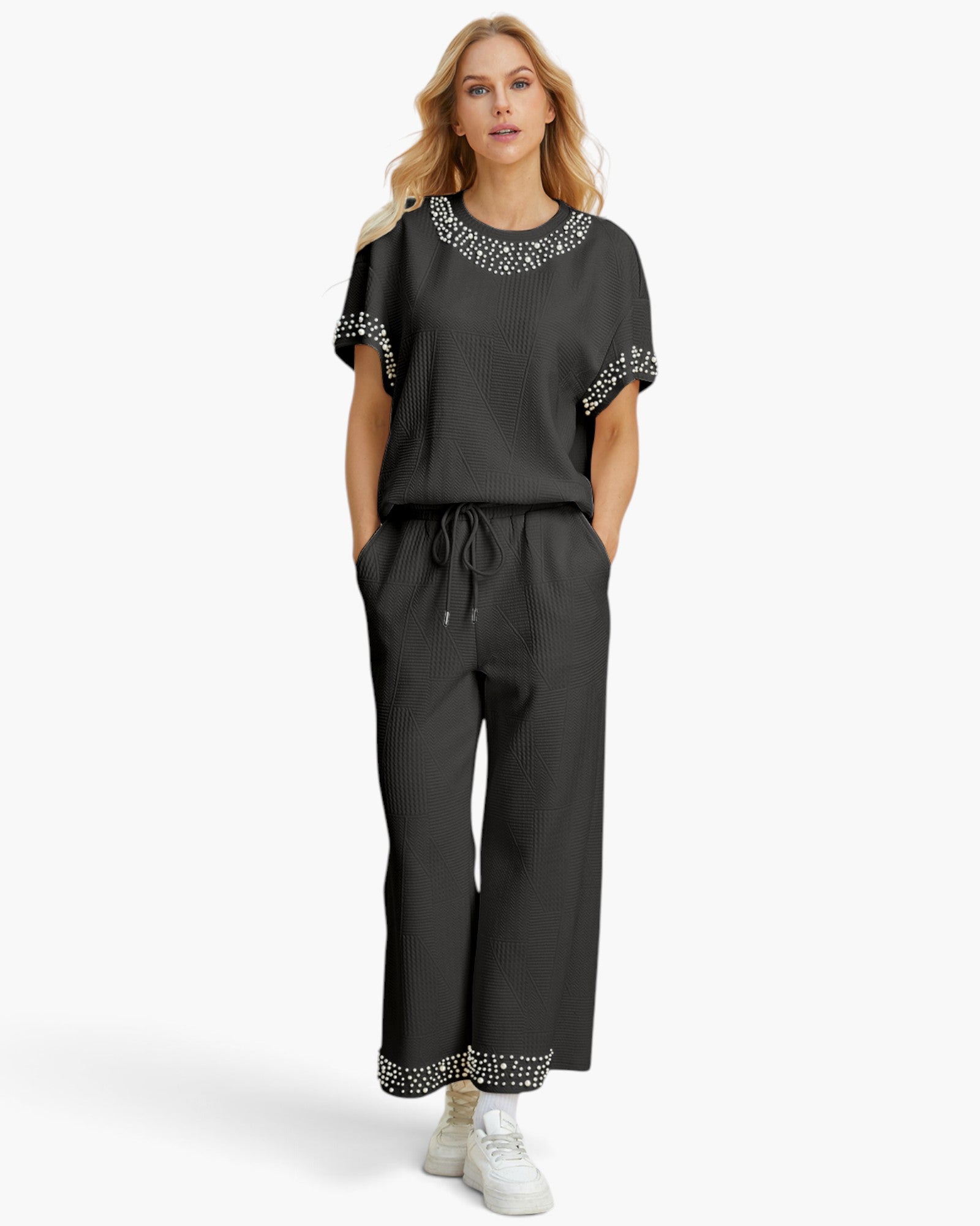 A woman modeling a dark gray two-piece set consisting of a short-sleeve top with silver rhinestone trim at the neckline and sleeve cuffs, and matching wide-leg trousers with matching rhinestone trim at the cuffs. She is wearing white sneakers against a plain white studio background.