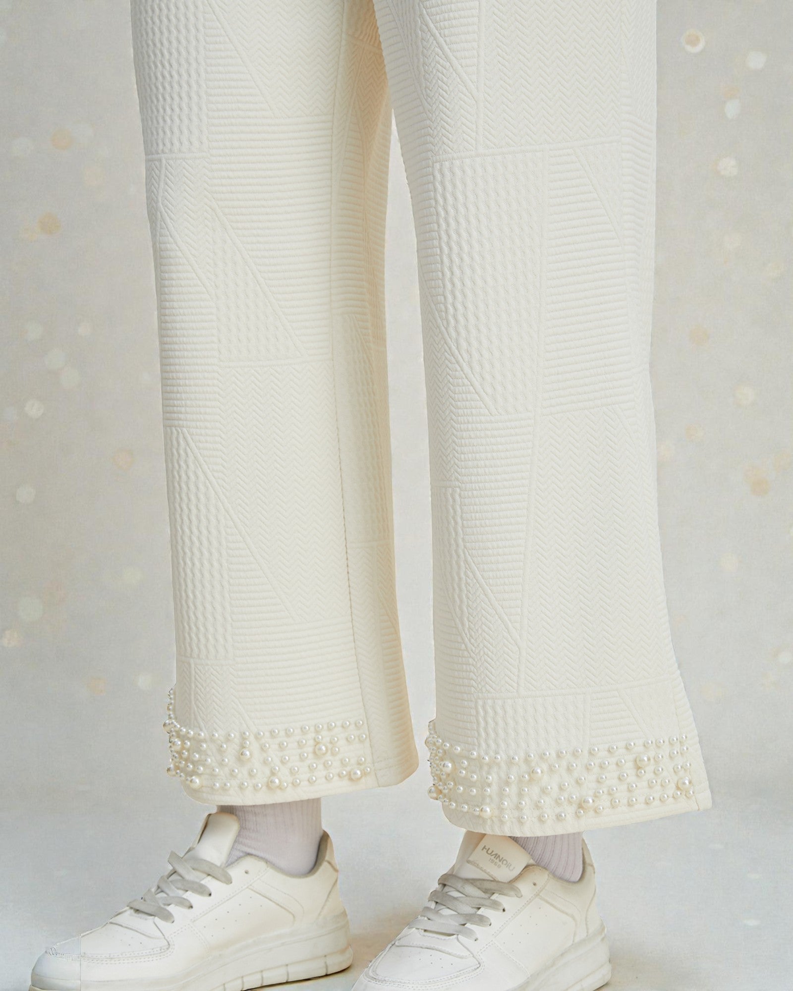 White textured pants with pearl details and white sneakers on a light gray background