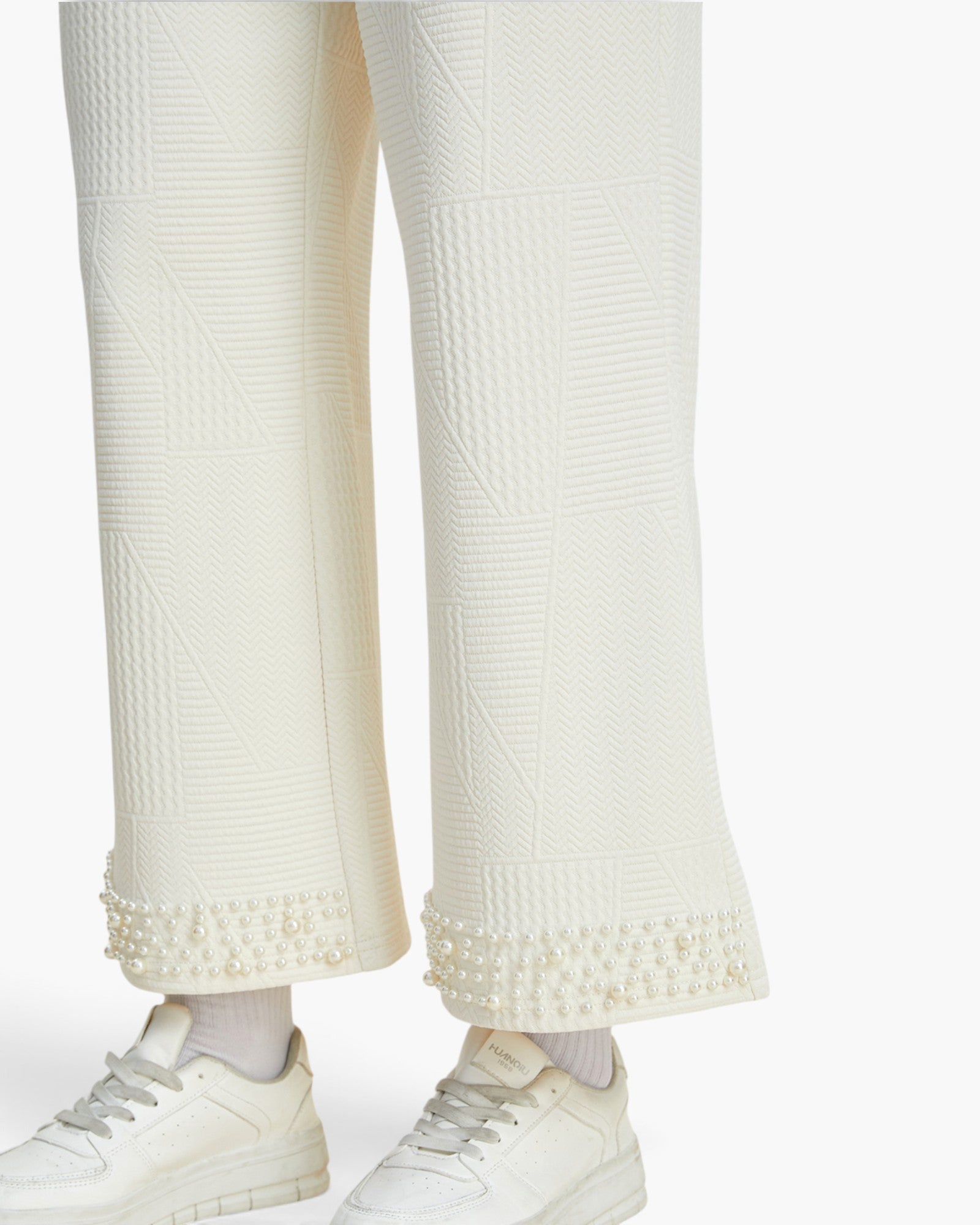Close-up of a person wearing textured off-white wide-leg trousers with pearl trim at the hem, paired with white sneakers and white socks, against a plain light background.