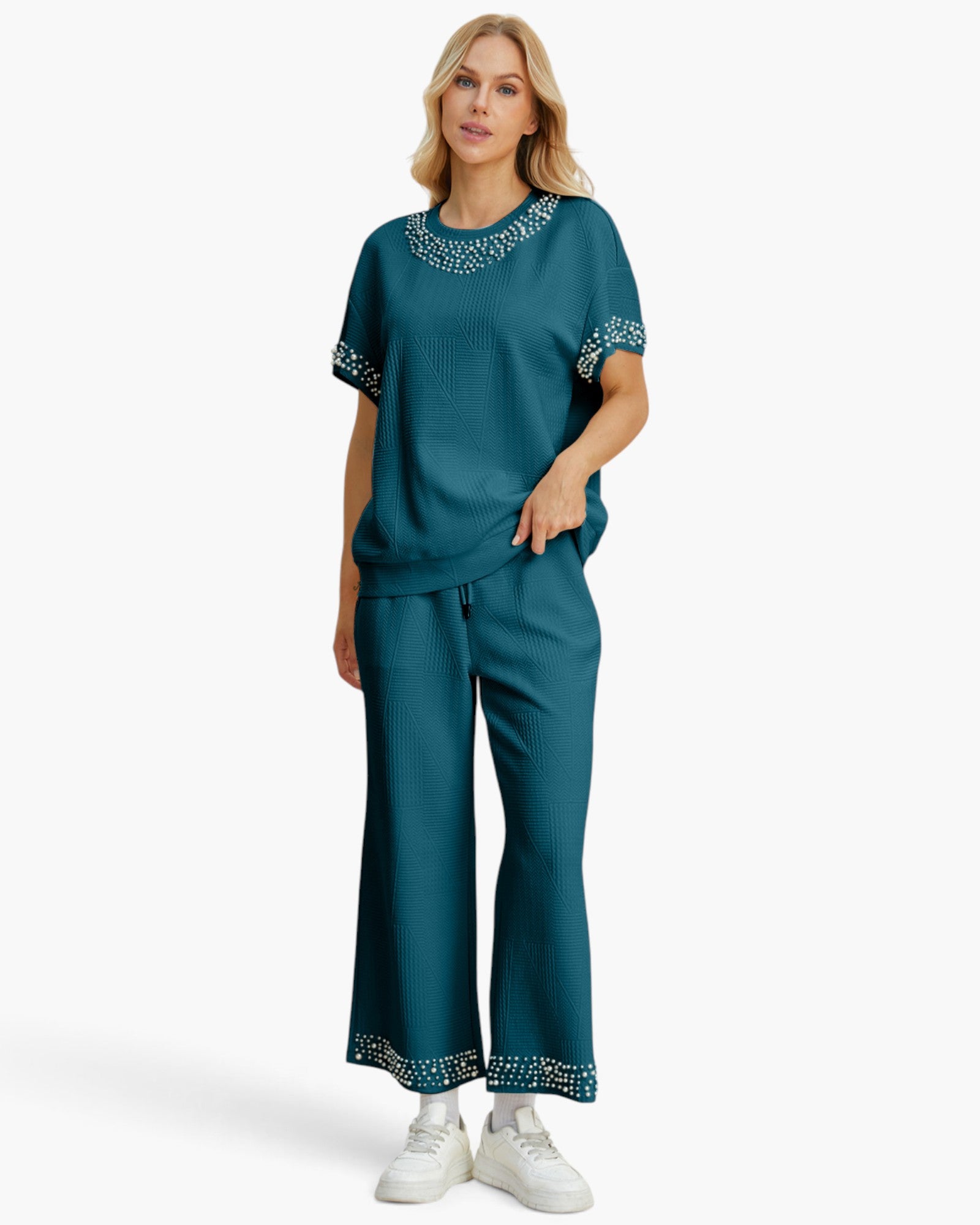 A woman modeling a matching teal lounge set consisting of a short-sleeve top with decorative silver embellishments around the neckline and sleeve cuffs, and matching wide-leg pants with embellished hems, paired with white sneakers on a light gray background.