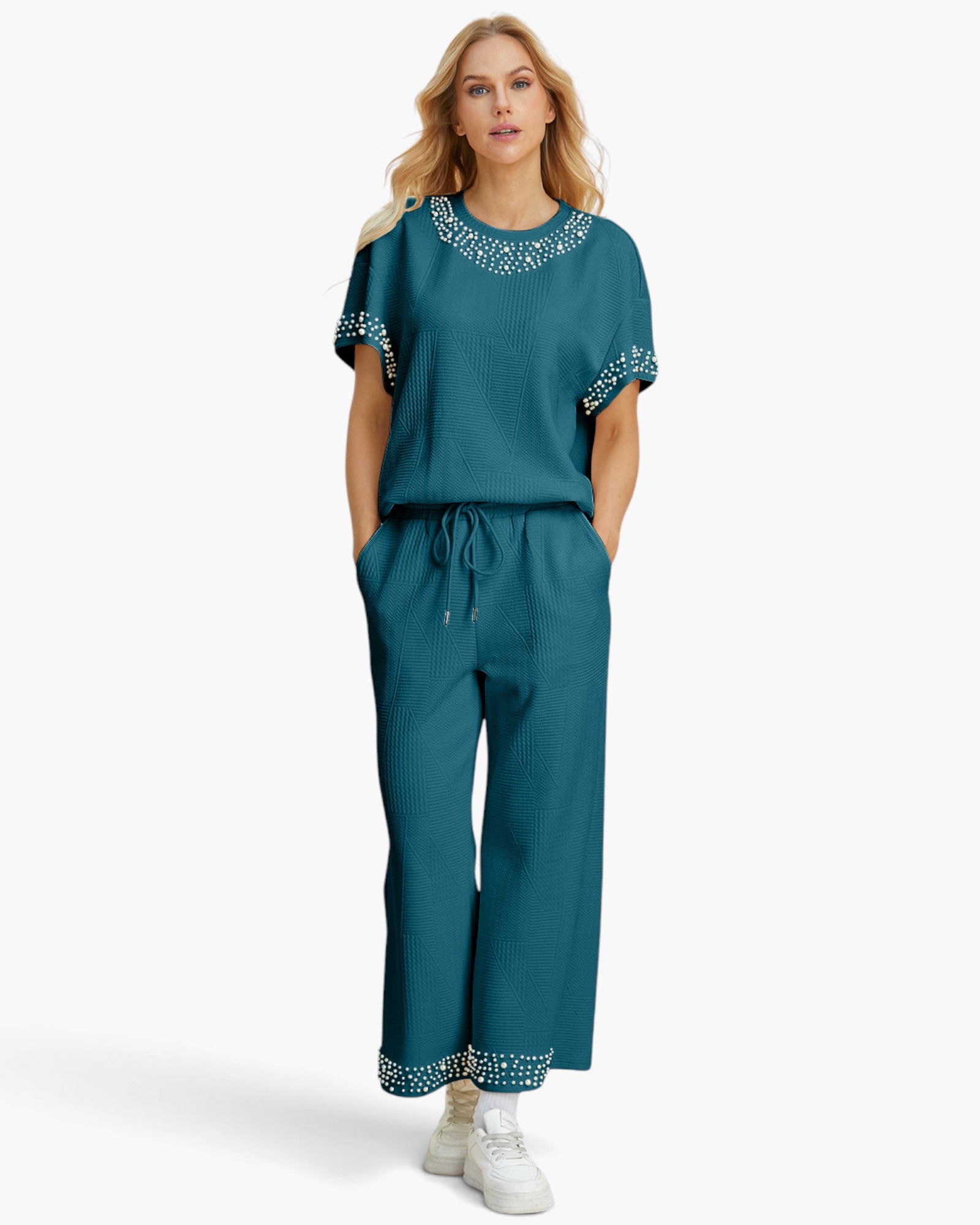 A woman modeling a matching teal two-piece lounge set consisting of a short-sleeve top with embellished yoke and sleeve cuffs, and matching wide-leg drawstring pants with embellished cuffs, paired with white sneakers against a light gray studio background.