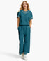 A woman modeling a matching teal two-piece lounge set consisting of a short-sleeve top with embellished yoke and sleeve cuffs, and matching wide-leg drawstring pants with embellished cuffs, paired with white sneakers against a light gray studio background.