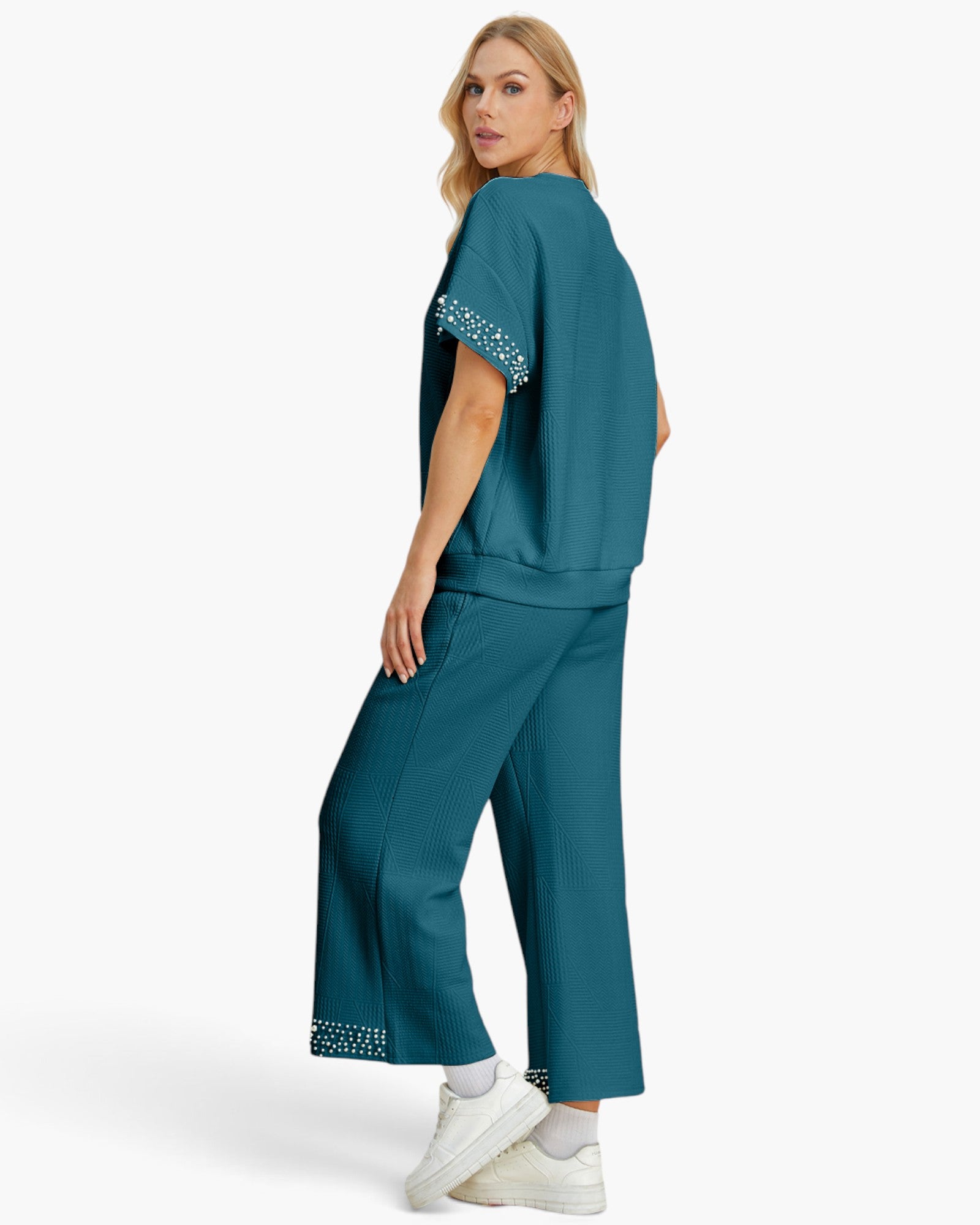 A woman modeling a matching teal lounge set consisting of a short-sleeve top with white beaded trim on the sleeve cuffs and matching wide-leg pants with beaded trim at the hem, paired with white sneakers against a light gray studio background.