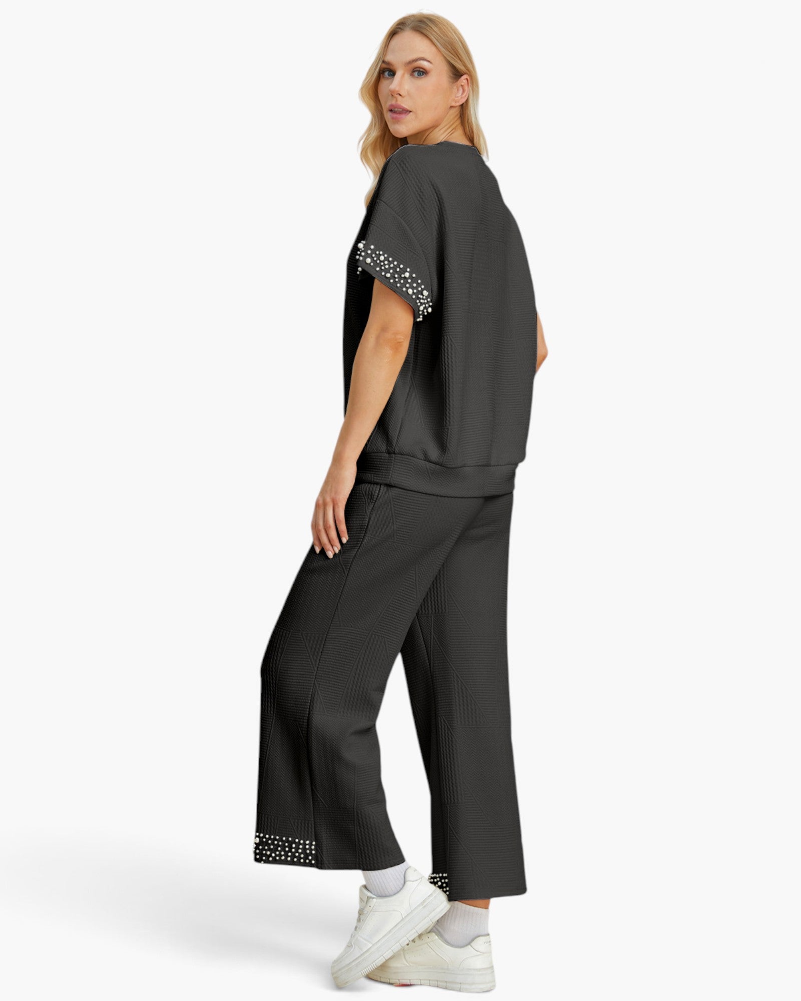 A woman modeling a dark gray two-piece lounge set consisting of a short-sleeve top with studded sleeve cuffs and matching wide-leg cropped pants with studded hem detailing, paired with white sneakers against a light gray studio background.