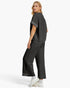 A woman modeling a dark gray two-piece lounge set consisting of a short-sleeve top with studded sleeve cuffs and matching wide-leg cropped pants with studded hem detailing, paired with white sneakers against a light gray studio background.