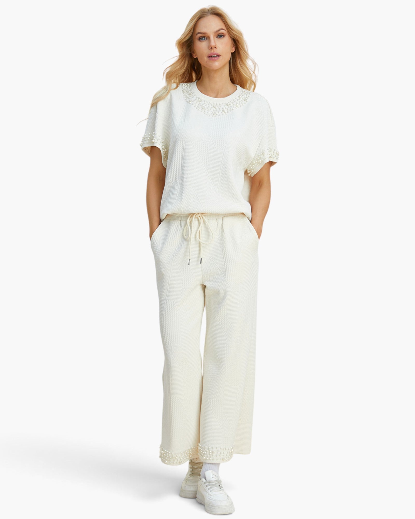 A woman modeling a matching cream-colored lounge set consisting of a short-sleeve top with lace trim and matching wide-leg drawstring pants, paired with white sneakers against a plain white background.
