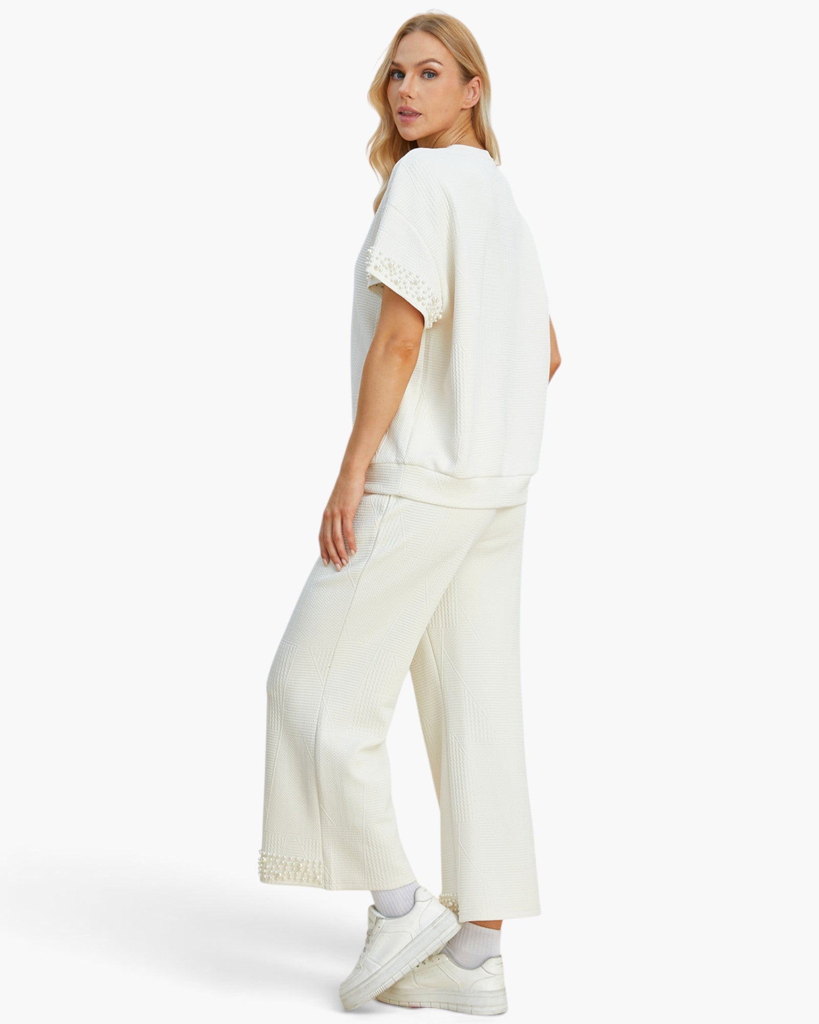A woman modeling a matching off-white lounge set consisting of a short-sleeve top with lace trim and wide-leg cropped pants, paired with white sneakers against a plain white studio background.