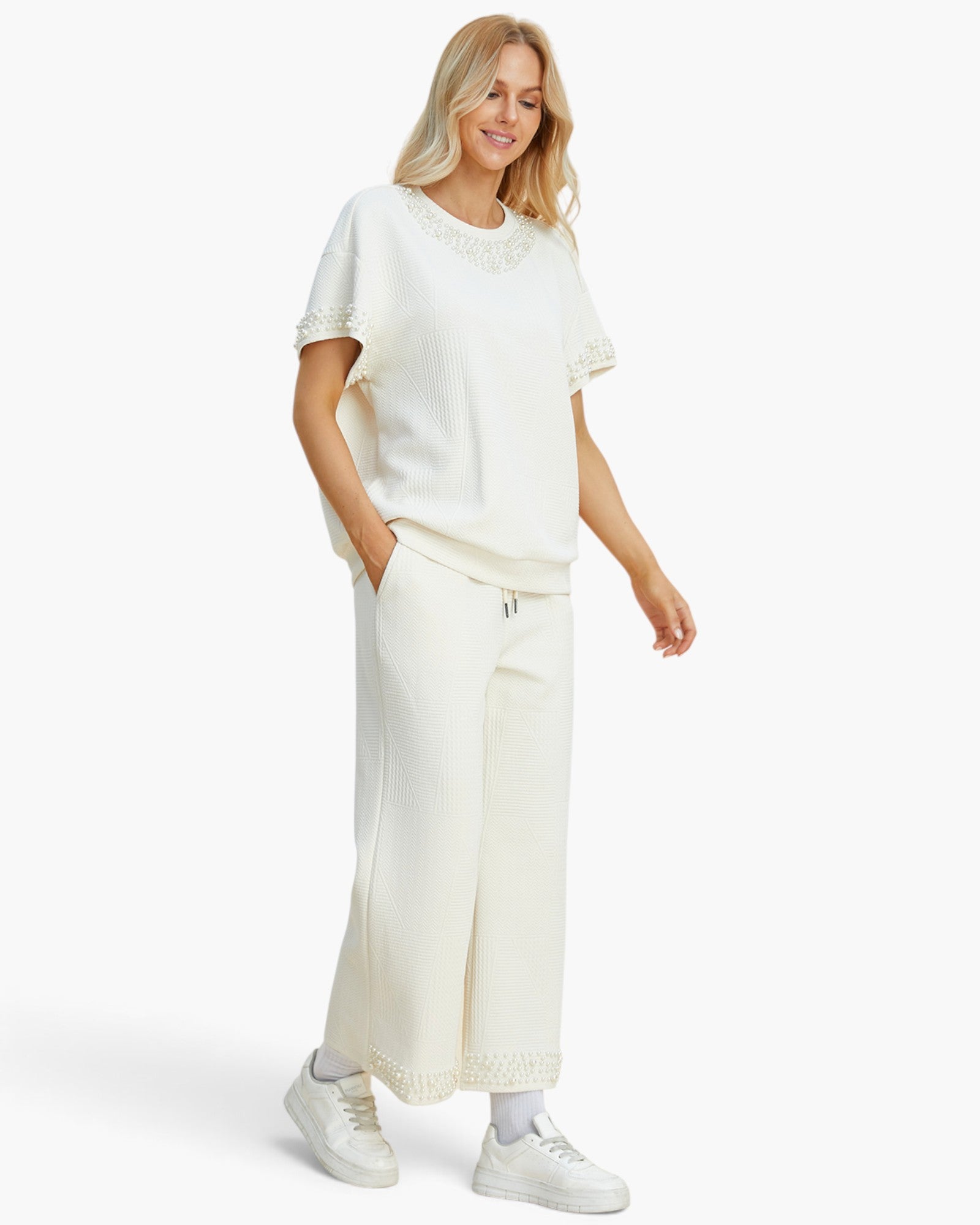 A woman modeling a matching cream-colored lounge set consisting of a short-sleeve top with lace trim and wide-leg cropped pants, paired with white sneakers, against a plain white studio background.