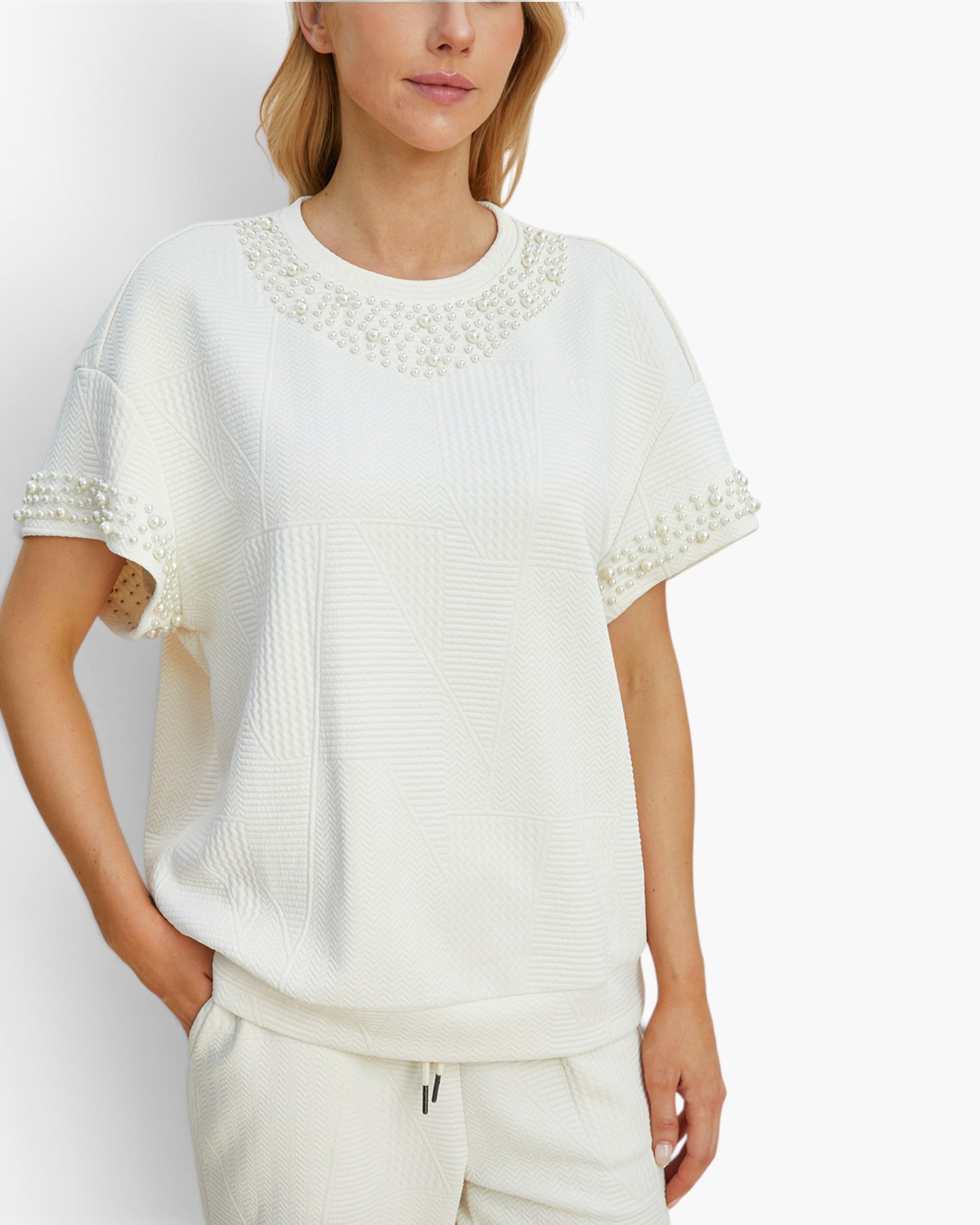 A woman wearing a white textured short-sleeve top with pearl embellishments around the crew neck and sleeve cuffs, paired with matching white pants, against a light gray studio background.