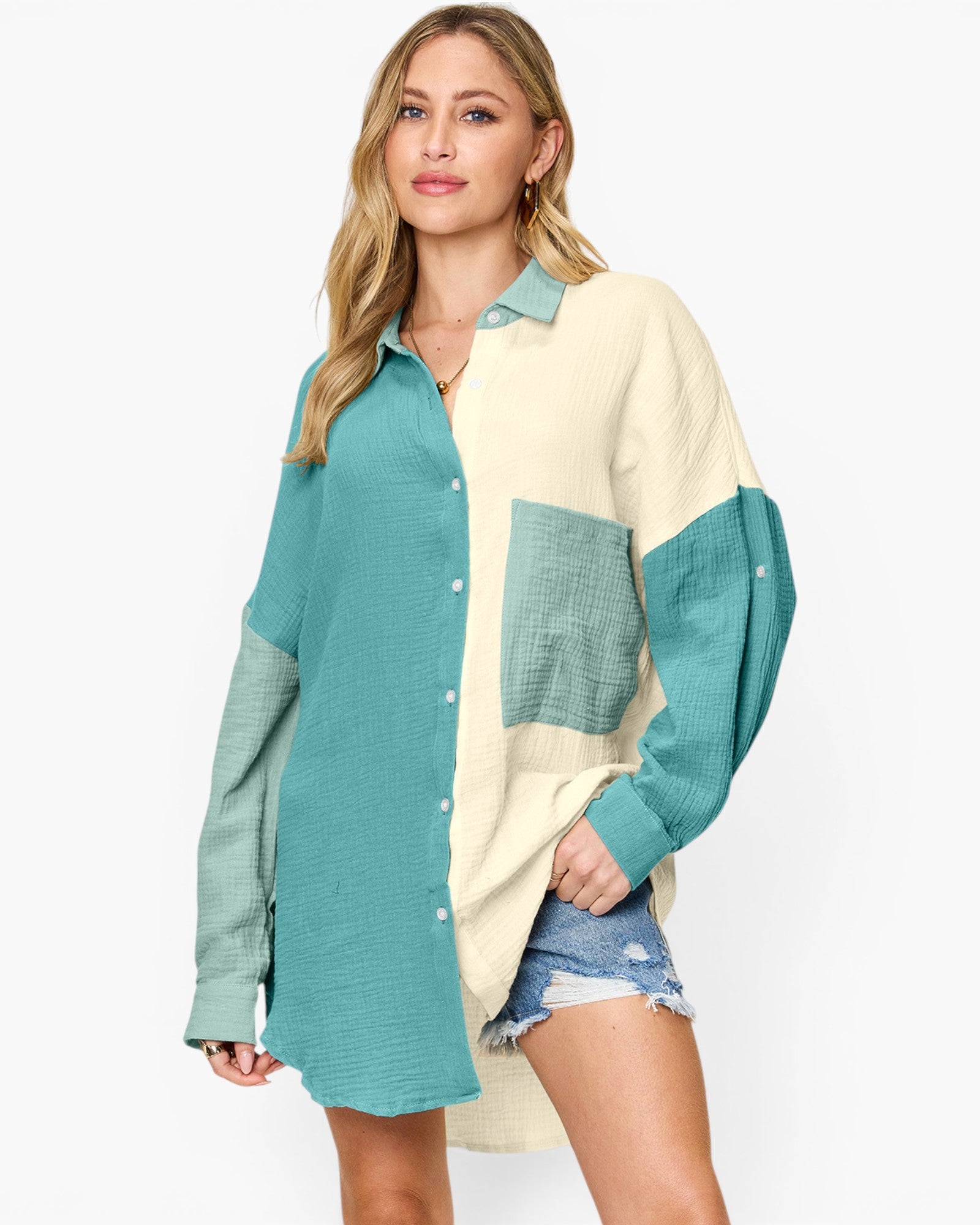 A woman modeling a long-sleeve colorblock button-up shirt with a teal green left side and sleeves, and a cream right side and chest panel, paired with denim shorts against a light gray studio background.