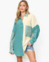 A woman modeling a long-sleeve colorblock button-up shirt with a teal green left side and sleeves, and a cream right side and chest panel, paired with denim shorts against a light gray studio background.