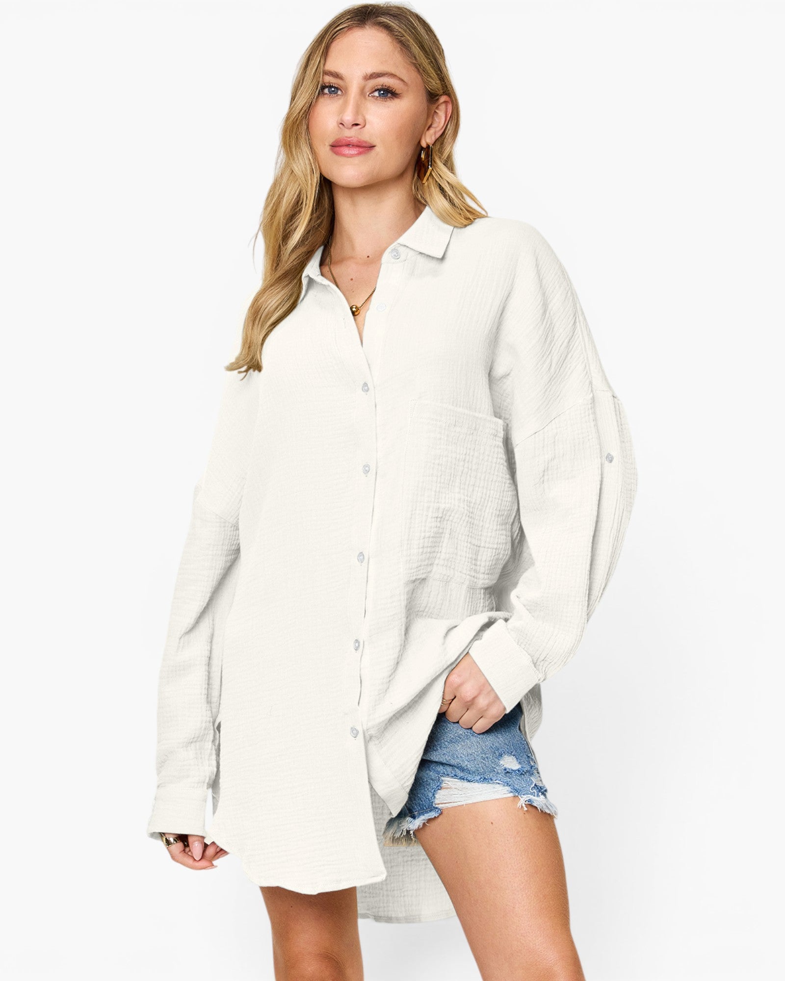 A woman modeling an oversized, long-sleeve button-up shirt in a textured off-white fabric, paired with distressed denim shorts against a plain white studio background.