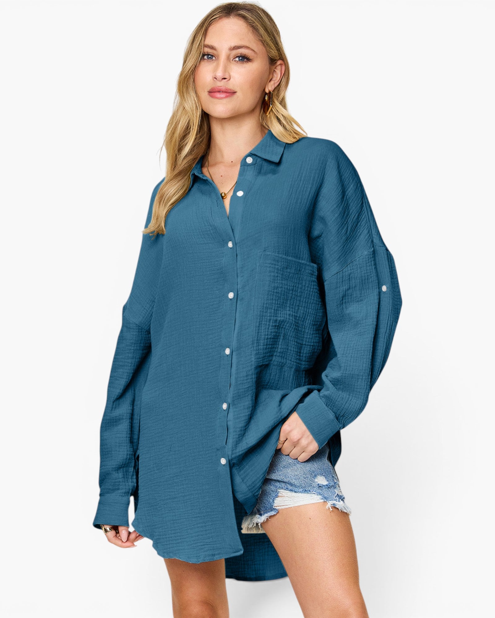 A woman modeling a teal blue textured button-up shirt dress with denim shorts against a plain white studio background.