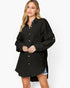 A woman modeling a black oversized button-down shirt dress with a textured crinkle fabric, long sleeves, and a collar, set against a plain white studio background.