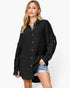 A woman modeling a black long-sleeve button-up shirt dress with a textured crinkle fabric, paired with distressed denim shorts, against a plain white studio background.
