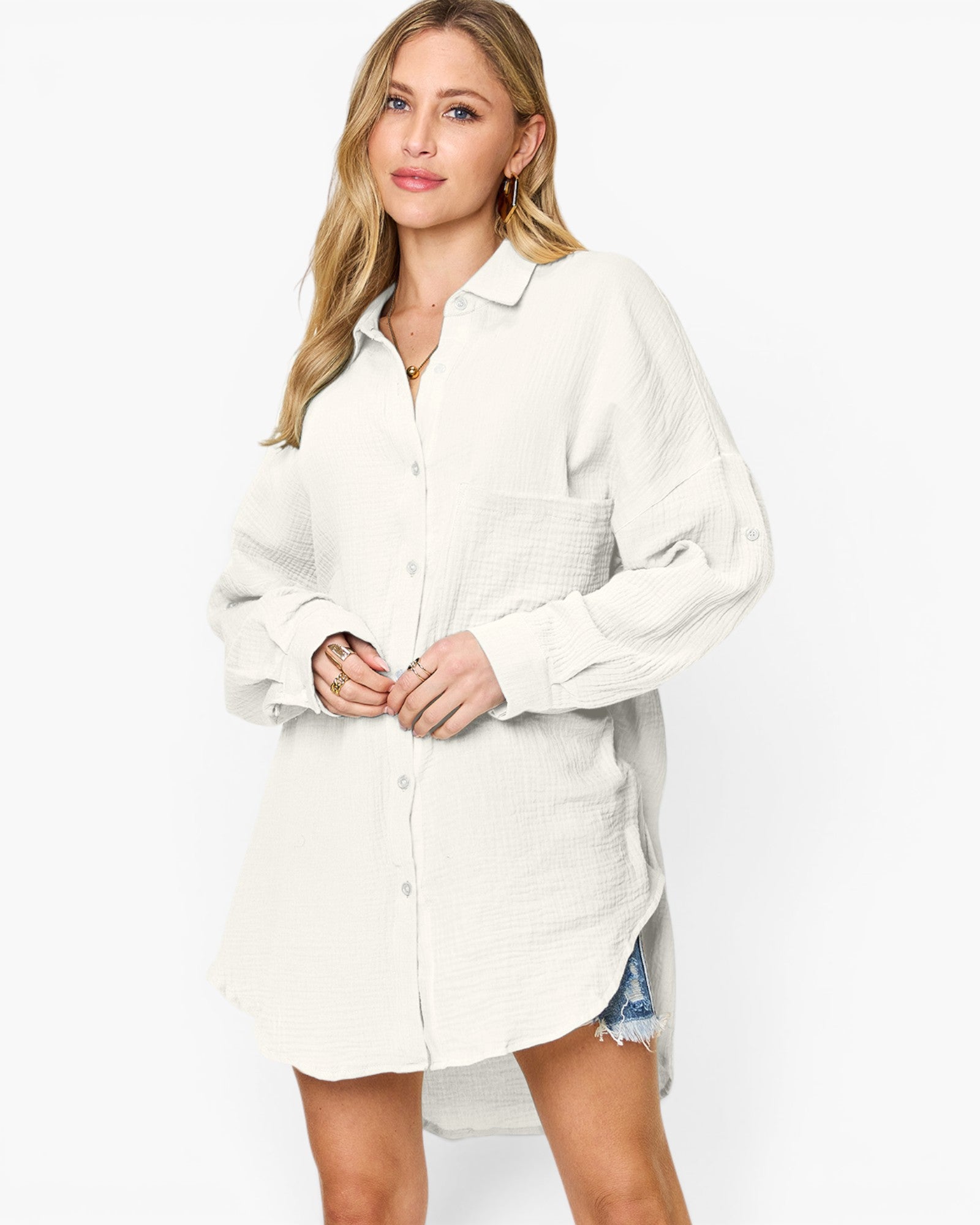 A woman modeling a white textured button-up shirt dress with a collar, long sleeves, and a high-low hem against a plain white studio background.