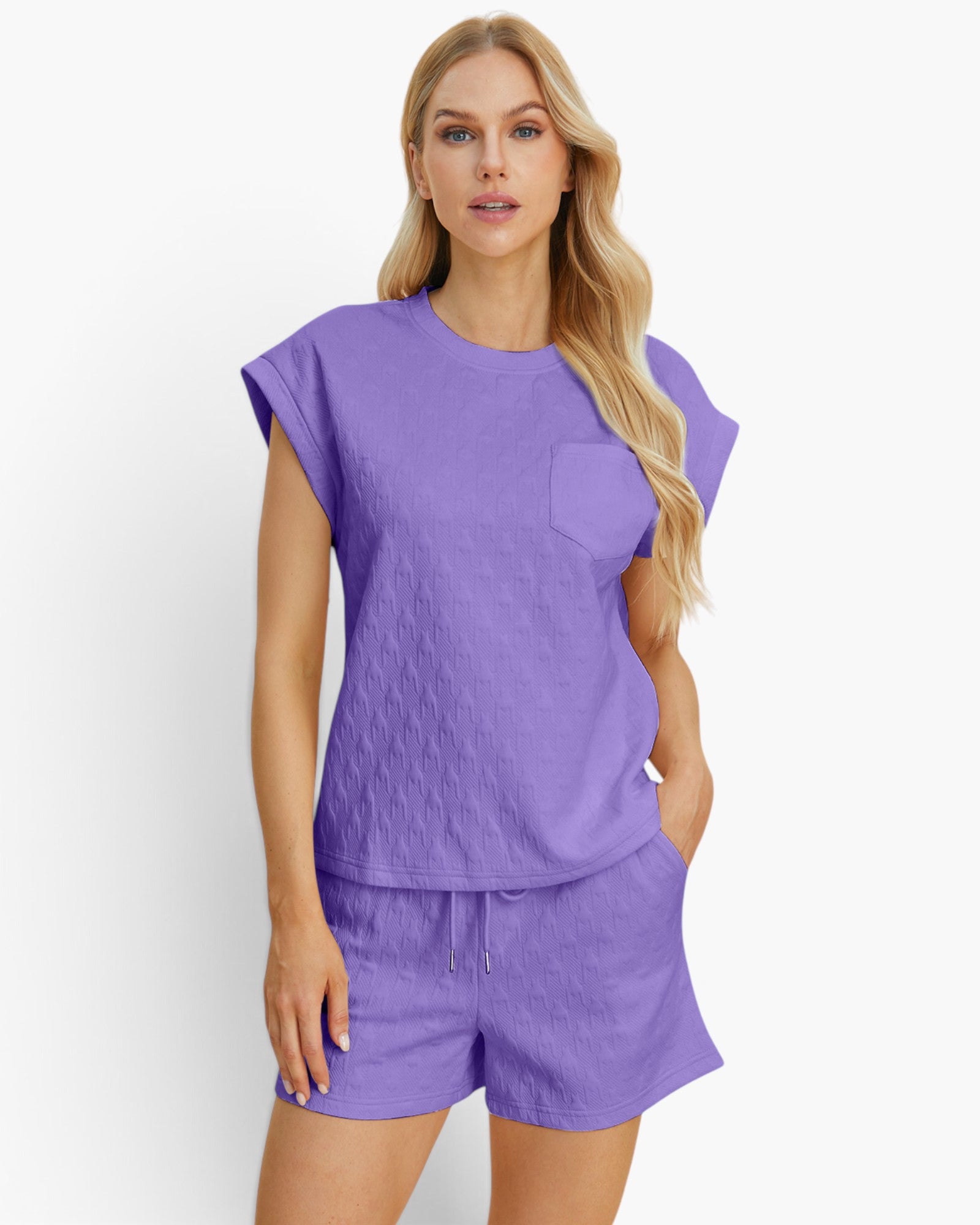 A woman modeling a matching lavender-purple lounge set consisting of a short-sleeve cropped top with a chest pocket and matching drawstring shorts, against a clean white studio background.