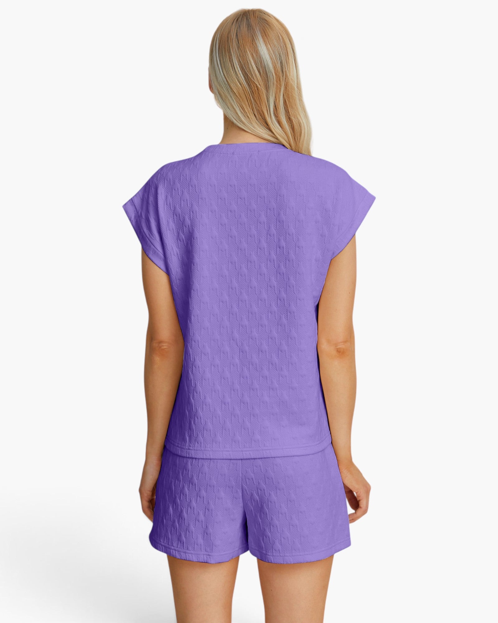 A woman viewed from the back wearing a matching lavender-purple textured lounge set consisting of a short-sleeve top and shorts, against a plain white studio background.