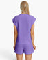 A woman viewed from the back wearing a matching lavender-purple textured lounge set consisting of a short-sleeve top and shorts, against a plain white studio background.