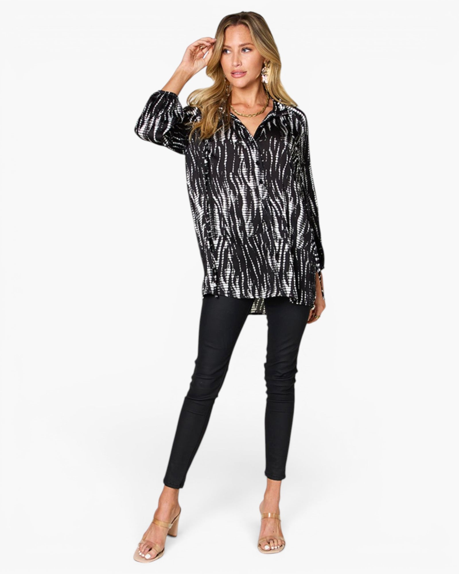 A woman modeling a black and white abstract patterned long-sleeve button-up shirt, paired with black skinny pants and clear high-heeled sandals, against a plain white background.