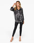 A woman modeling a black and white abstract patterned long-sleeve button-up shirt, paired with black skinny pants and clear high-heeled sandals, against a plain white background.