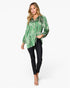 A woman modeling a green and white abstract patterned long-sleeve blouse with black skinny pants and clear high-heeled sandals, photographed against a light gray studio background.