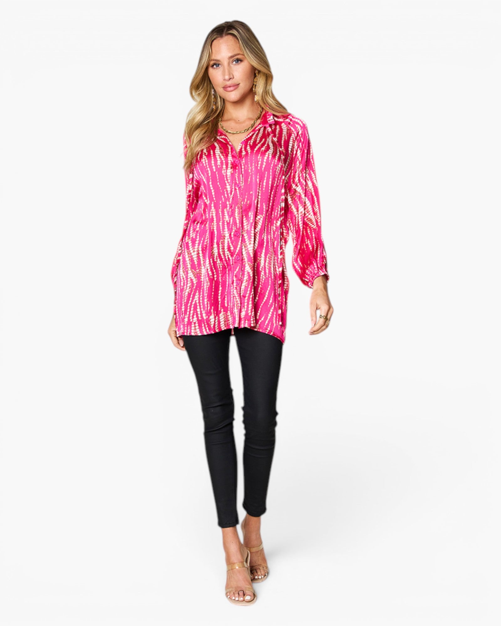 A woman modeling a vibrant pink and white abstract patterned long-sleeve blouse with black skinny pants and clear high-heeled sandals, set against a plain white studio background.