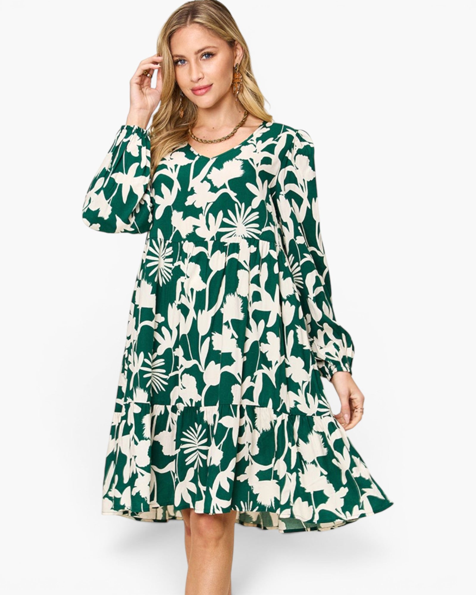 A woman modeling a long-sleeve knee-length dress with a bold green and off-white tropical floral print, set against a plain light gray studio background.