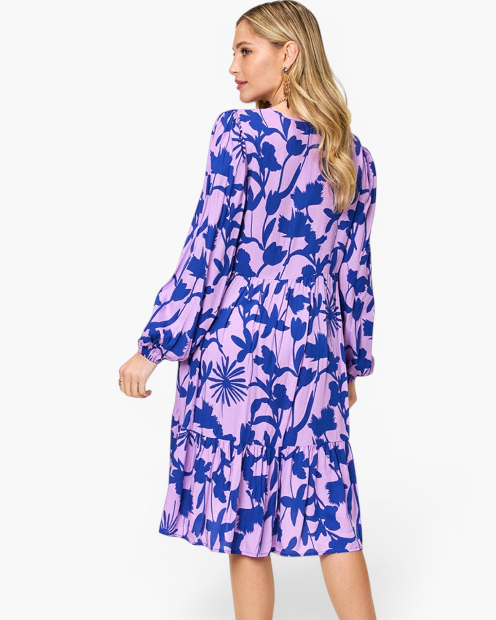 A woman modeling a long-sleeve midi dress with a purple and blue floral pattern, shown from a three-quarters back view against a light gray studio background.