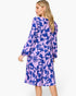 A woman modeling a long-sleeve midi dress with a purple and blue floral pattern, shown from a three-quarters back view against a light gray studio background.